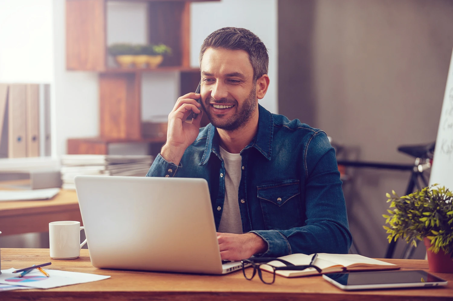 A man in a denim jacket speaks on his phone while working on a laptop at a wooden desk, surrounded by plants and office supplies.